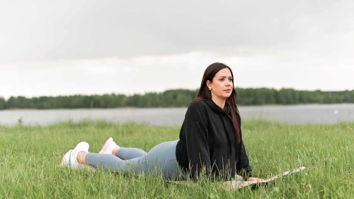 Woman lying on a yoga mat in a grassy field by a lake, promoting relaxation and wellness.